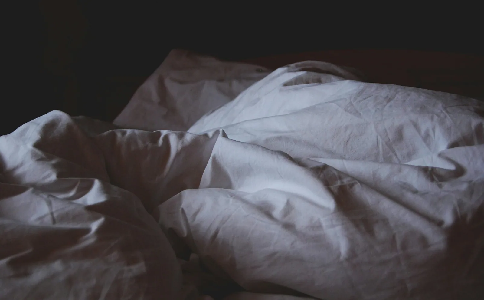 A rumpled white duvet on an empty bed in the dark