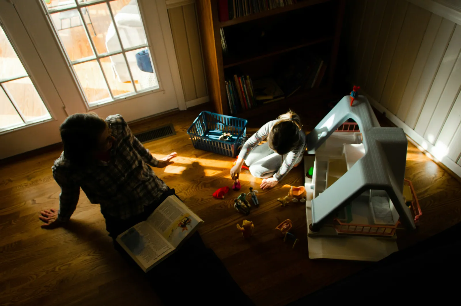 A parent reading in an armchair by a window while their child plays alone on the floor nearby