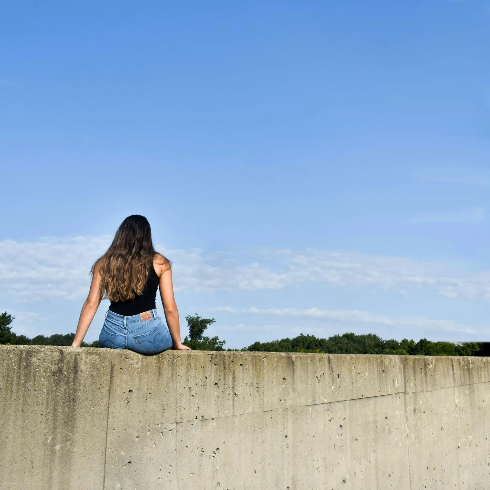 A teenage girl sitting alone on a low wall beneath a wide blue sky, looking out across the landscape