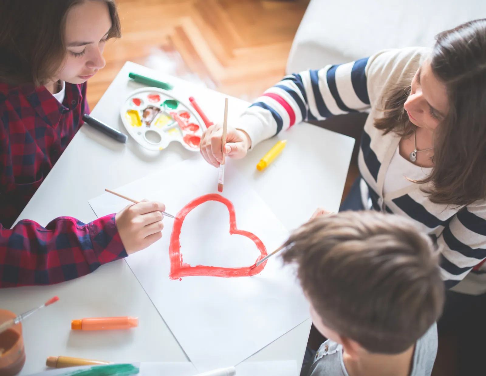 A parent and two children painting a large red heart together at a kitchen table