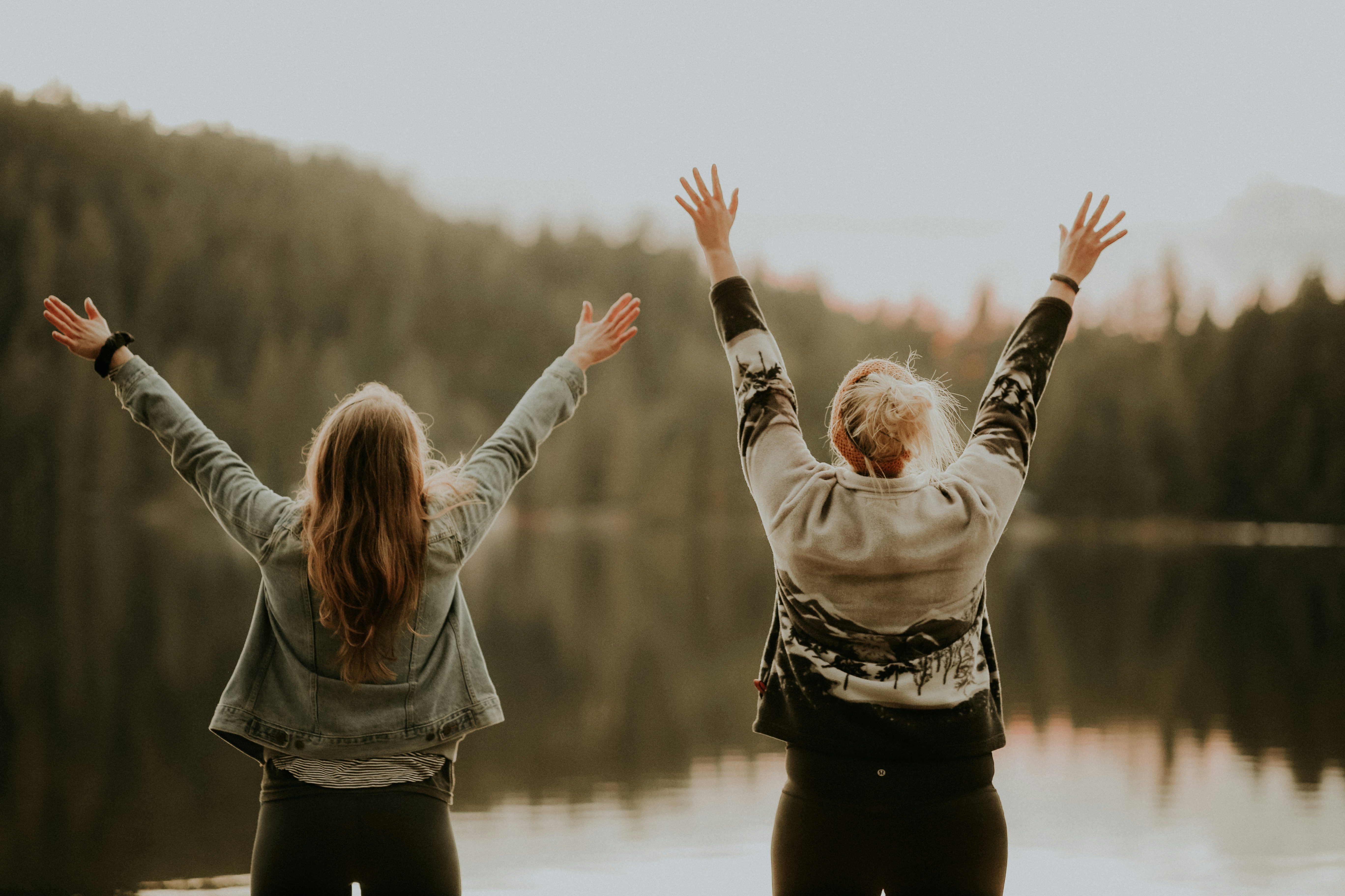 Two teenagers standing by a lake with their arms raised towards the hills