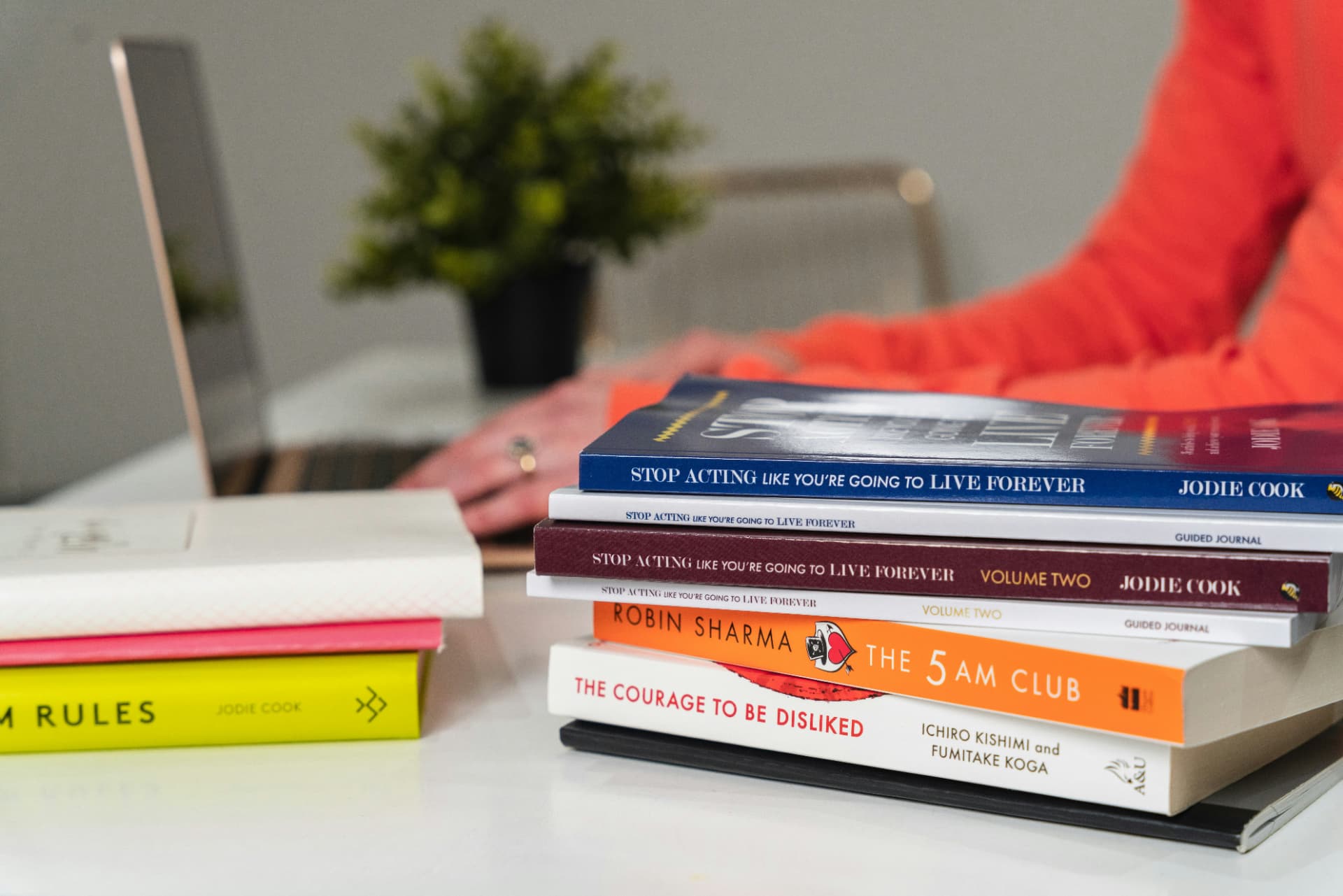 A stack of books on a desk with a laptop and morning light