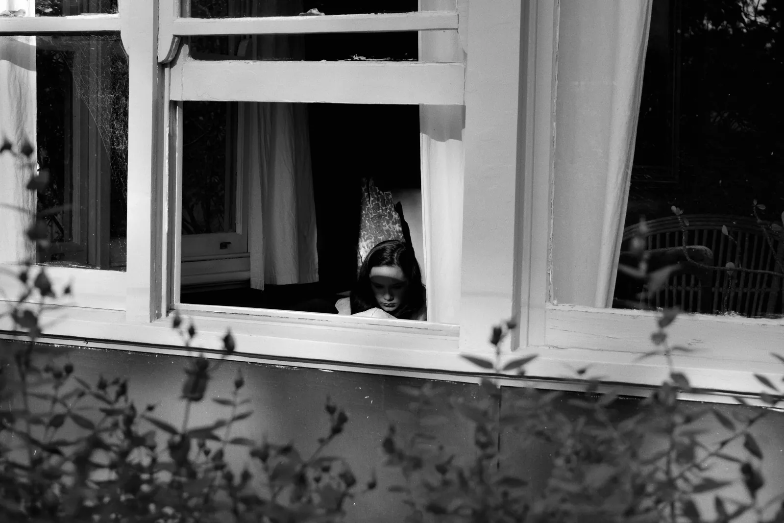A black and white photograph of a child looking out through a bedroom window