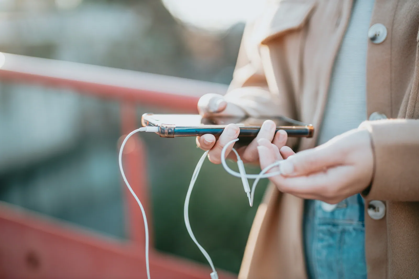 A teenager plugging earphones into a phone while standing outside, half turned away