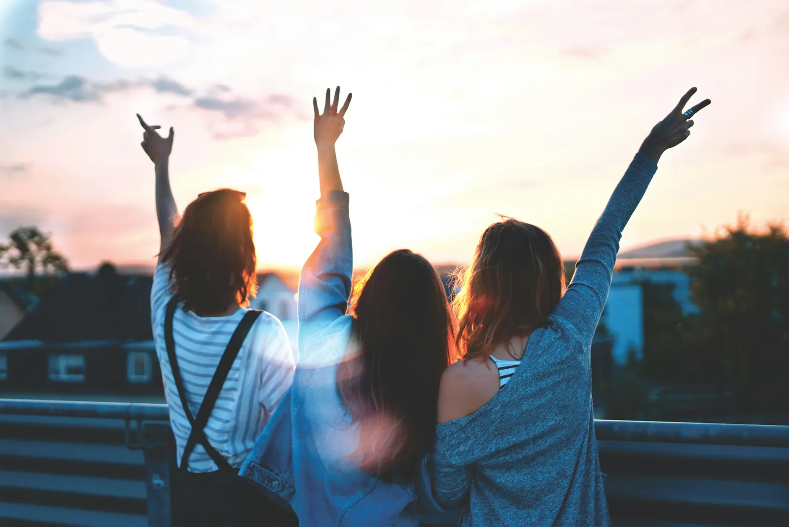 Three teenagers standing together at sunset with their arms raised, seen from behind