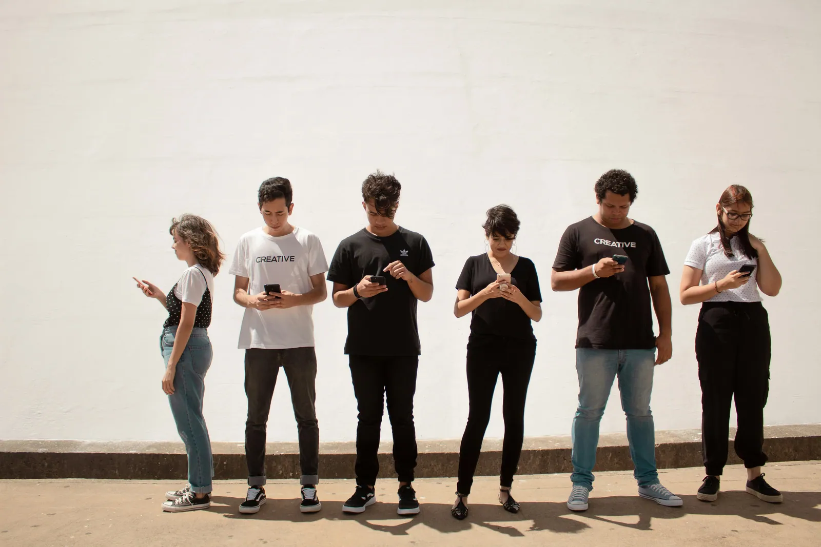 A row of teenagers standing against a white wall, every one of them looking down at a phone