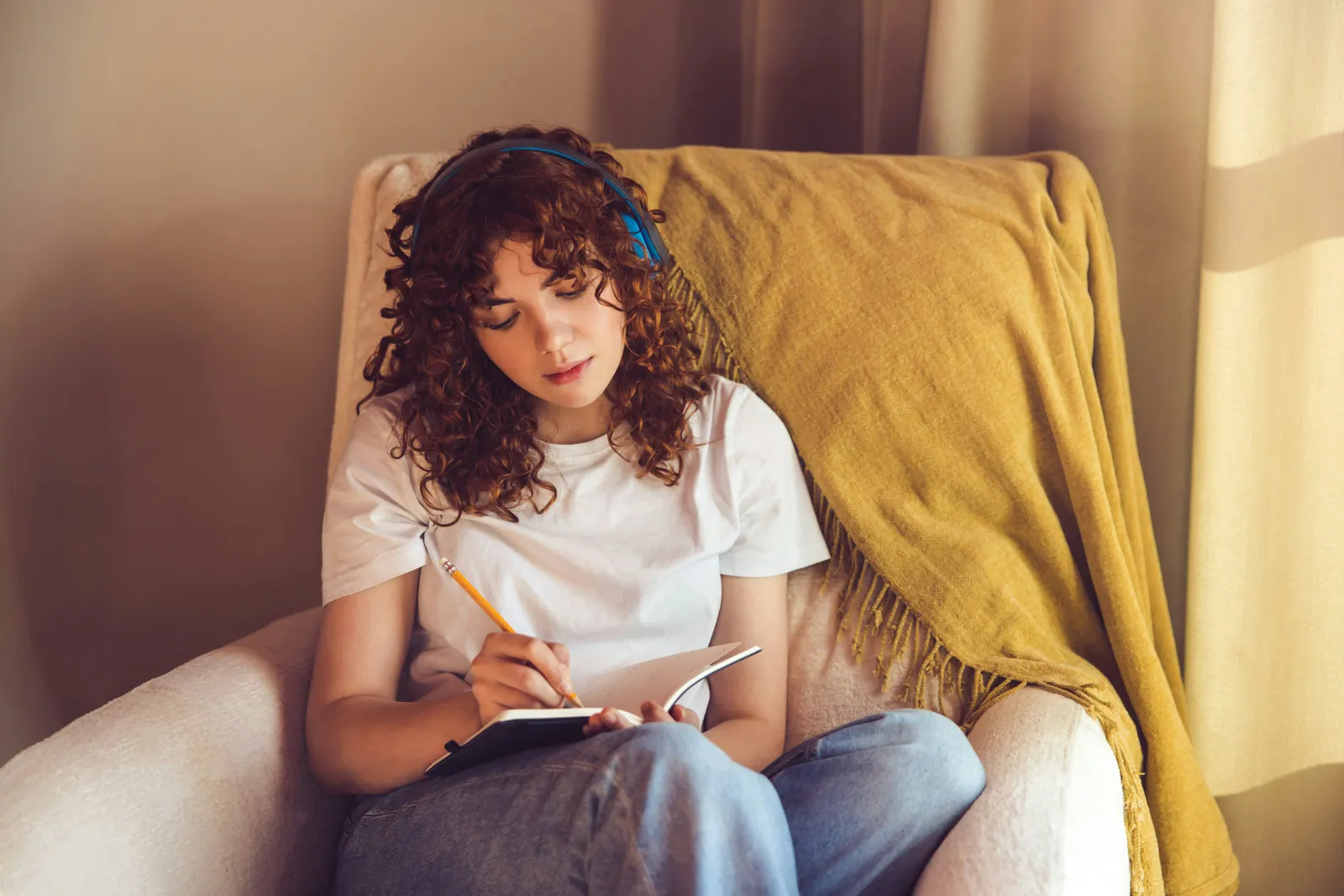 A teenage girl wearing headphones and writing reflectively in a notebook in a sunlit armchair