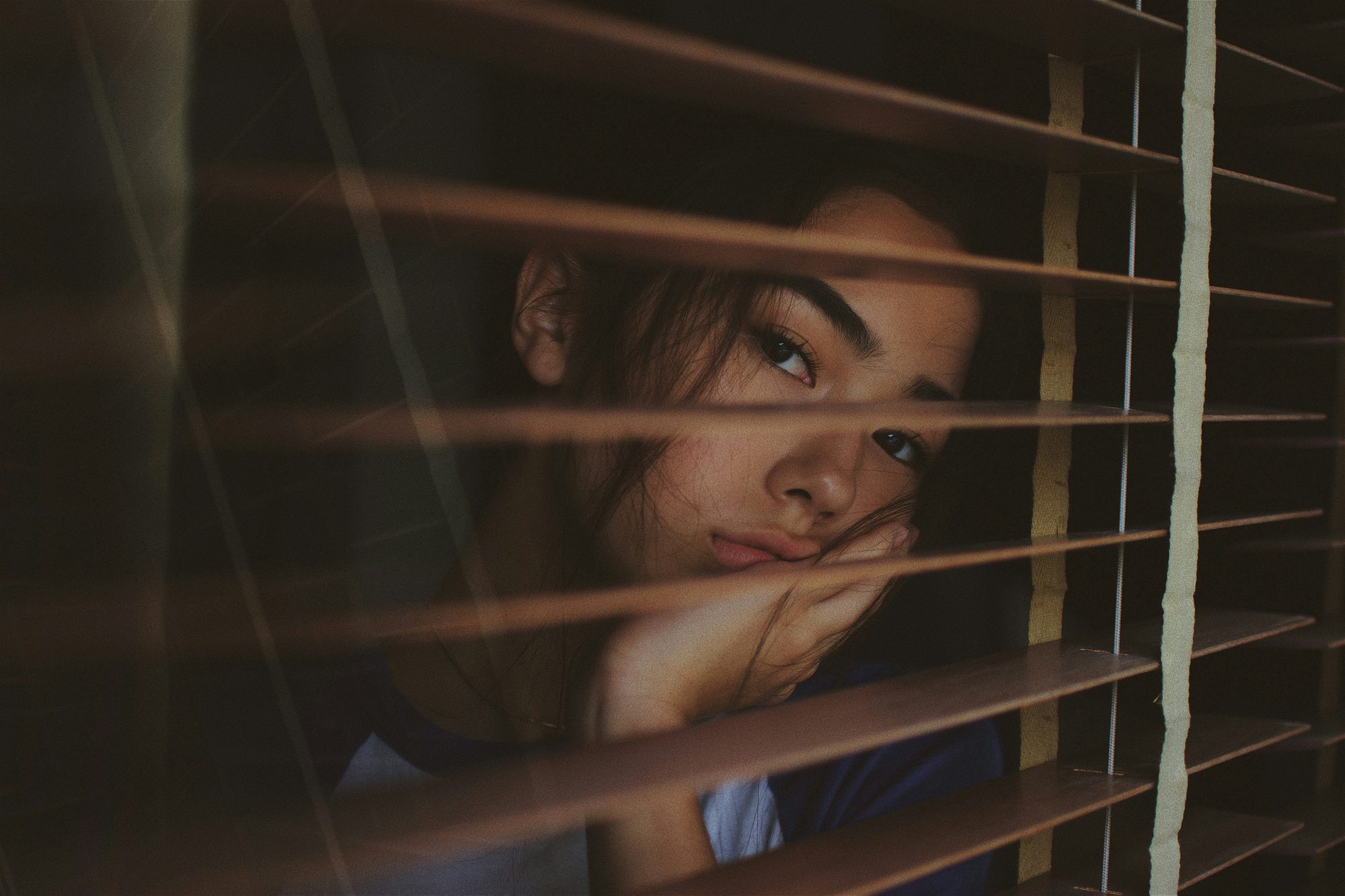 A teenager looking out through half-open window blinds in warm light