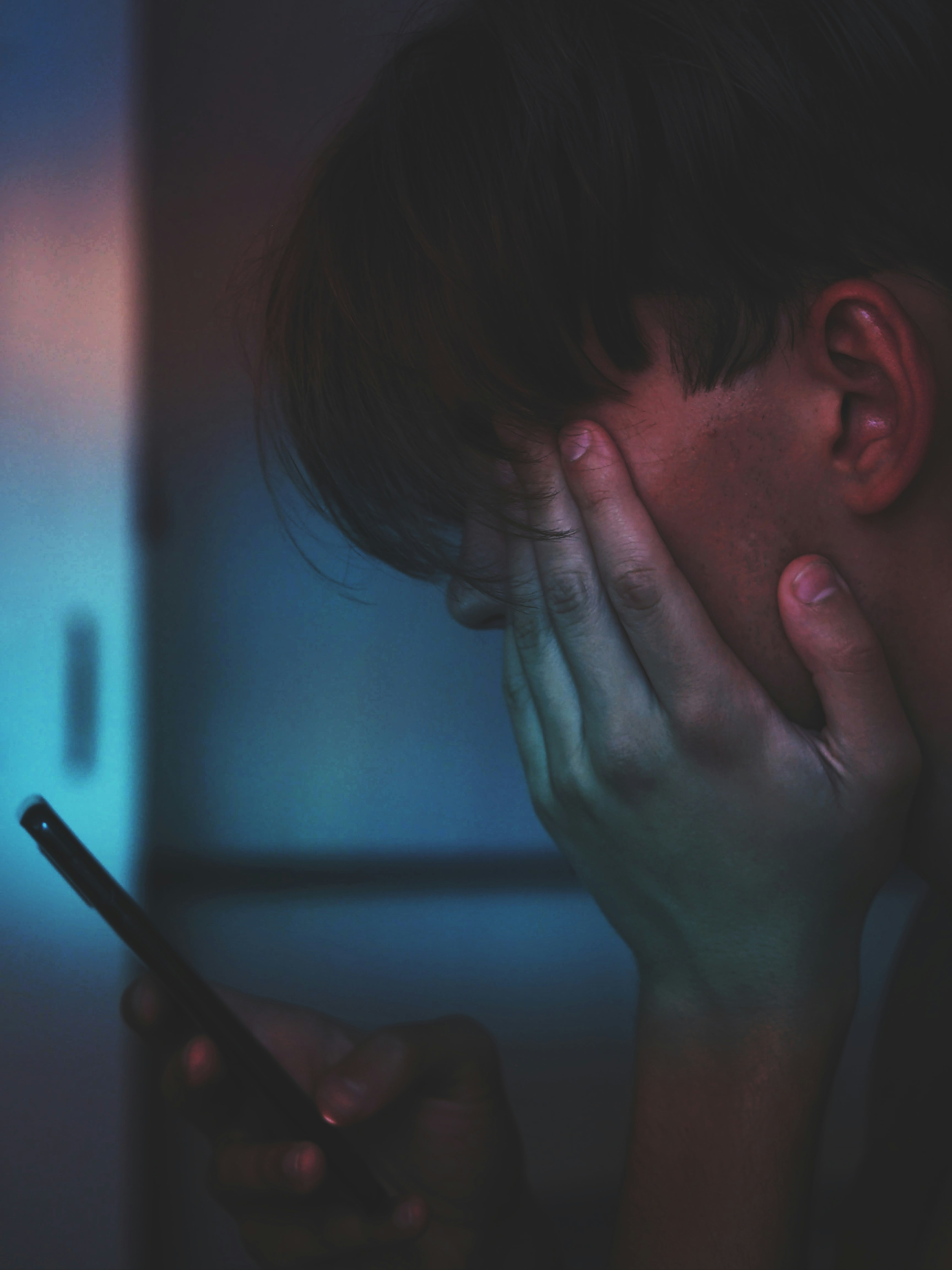 A teenager sitting in the dark with their face in their hands, lit only by a phone screen