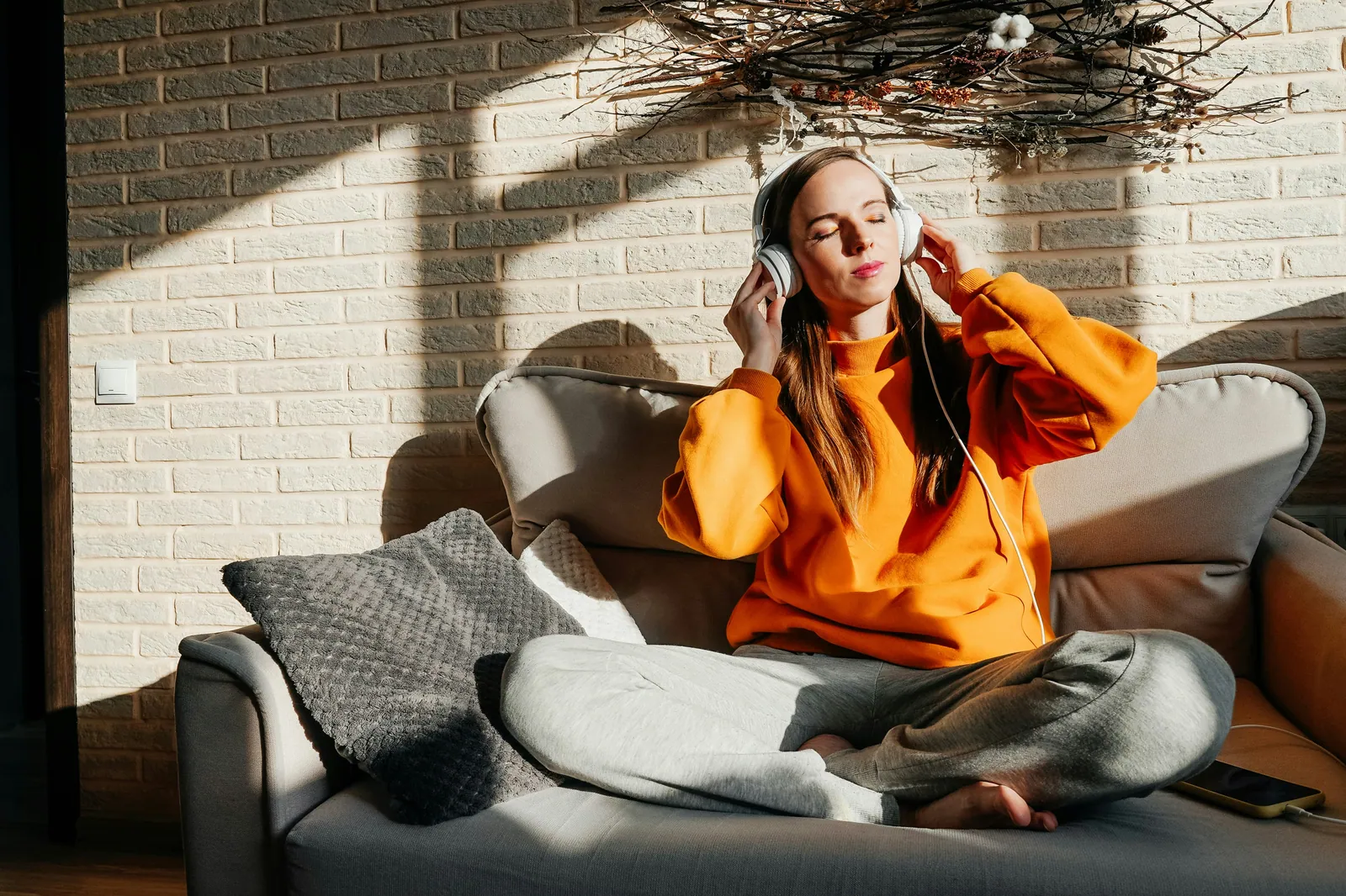 A woman sitting cross-legged on a sofa with headphones on and her eyes closed
