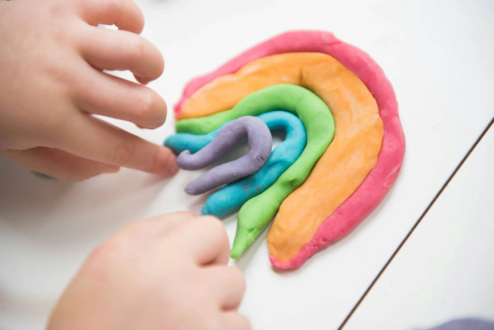 A child's hands shaping a rainbow from coloured modelling dough on a pale table