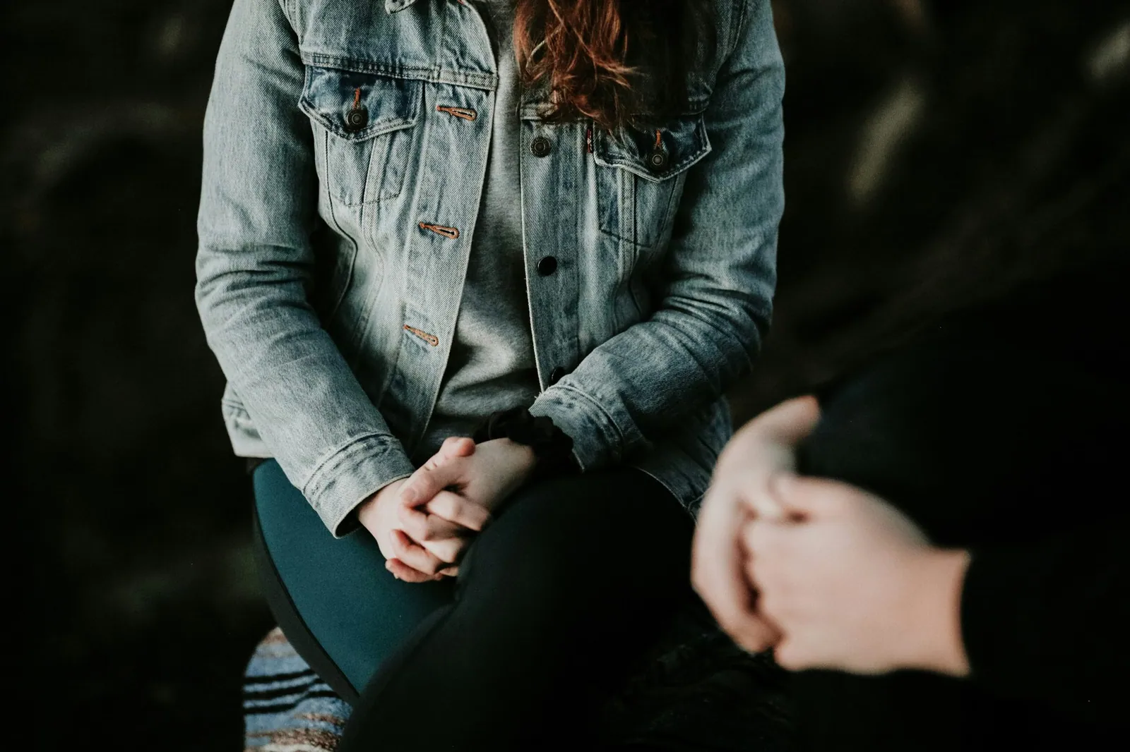 A teenager in a denim jacket sitting with hands clasped in her lap, shoulders drawn in