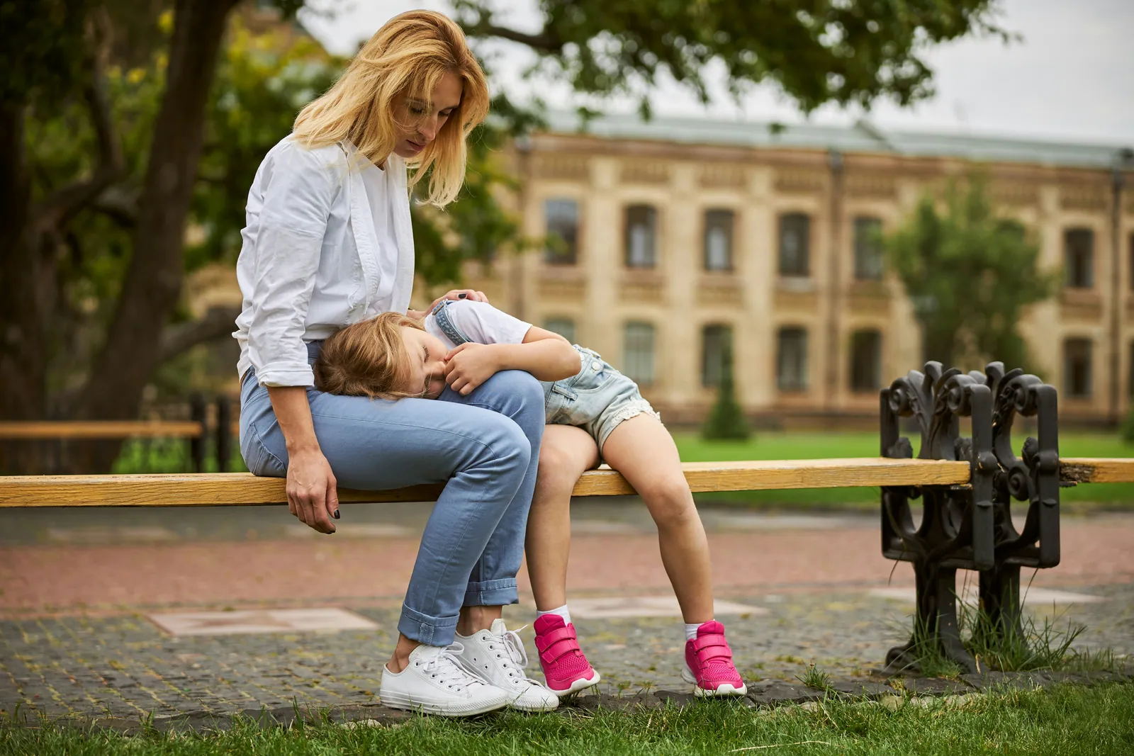 A child collapsed across a parent lap on a park bench after school, still in their trainers