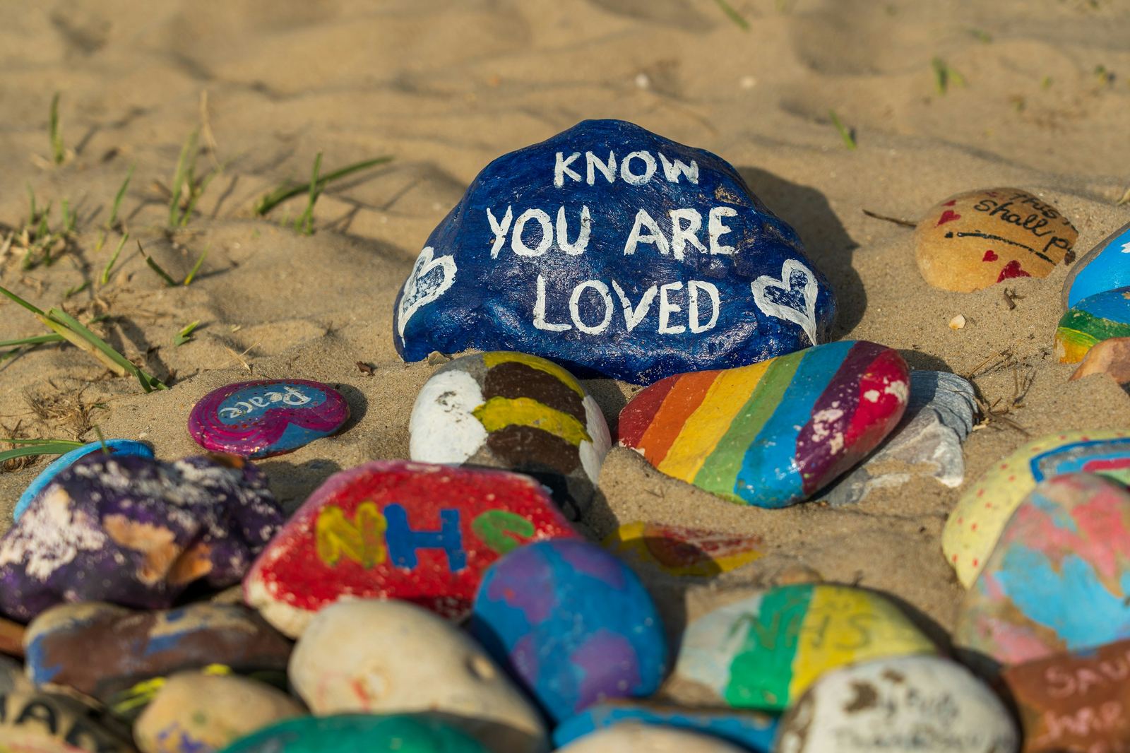 Painted rocks on sand, one reading know you are loved beside a rainbow