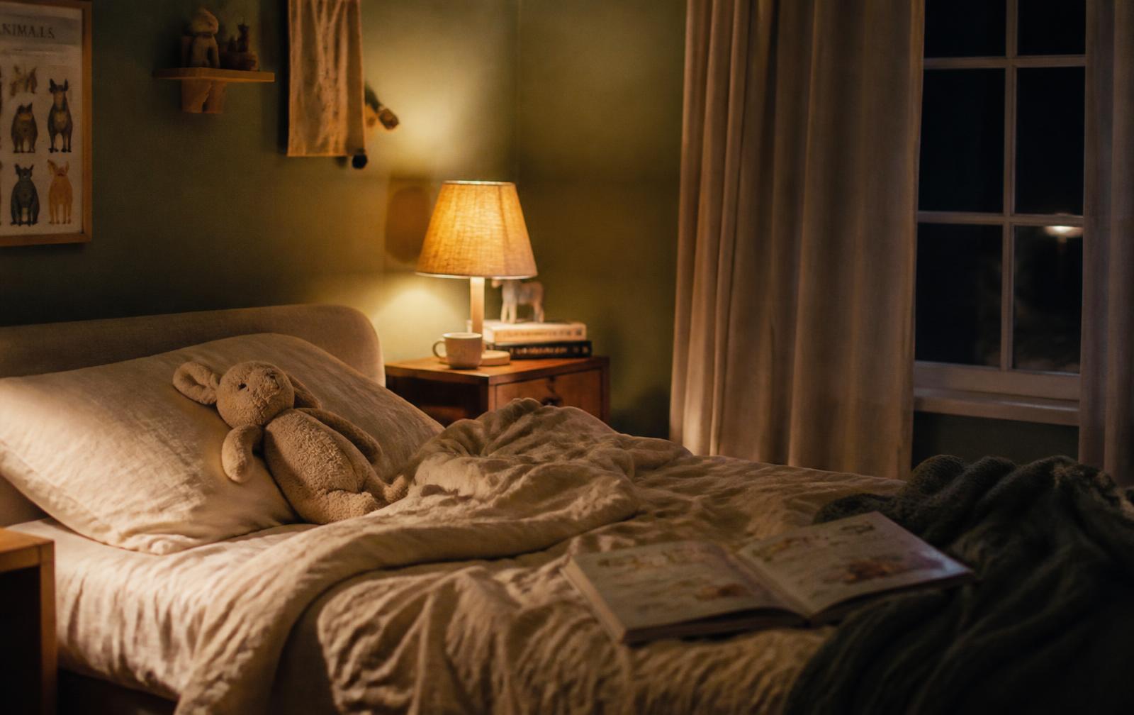 A child's bedroom at night with a soft toy, an open book and a lamp still glowing