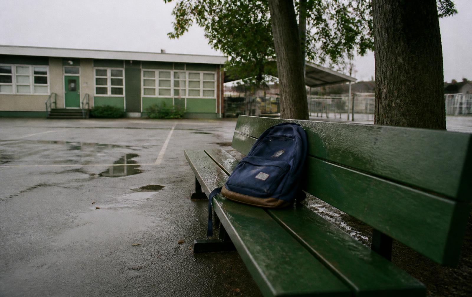 An empty green school bench under overcast light with a single backpack left behind