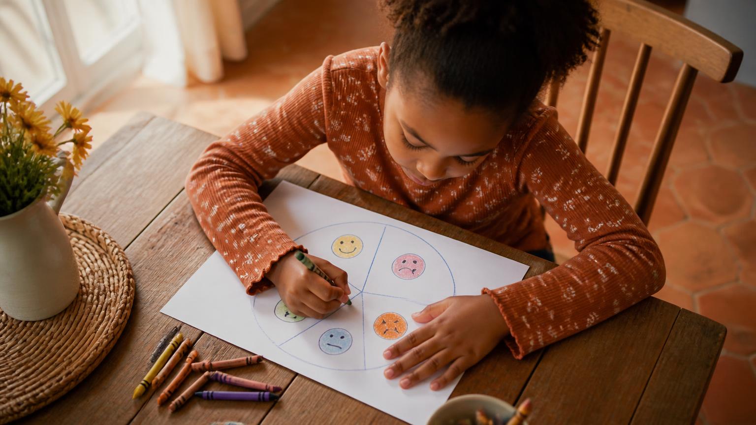 A young girl at a kitchen table colouring in a hand-drawn feelings wheel with crayons