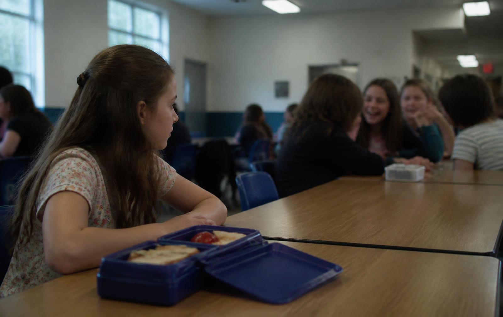 A girl sitting alone with her lunchbox at the end of a school hall table while classmates laugh together further along