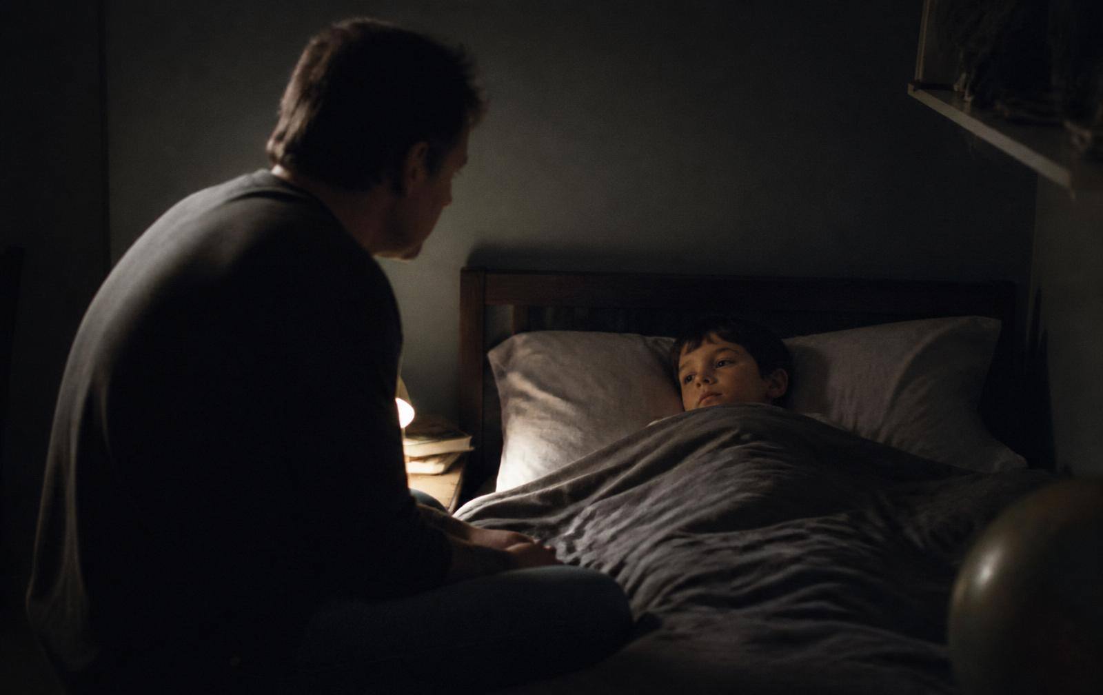 A parent sitting on the edge of a child's bed in a dim bedroom at night