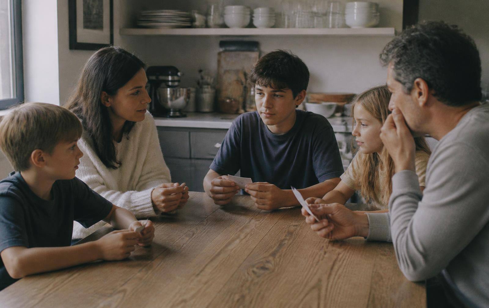 A family of five around a kitchen table, each holding a small slip of paper, mid-discussion