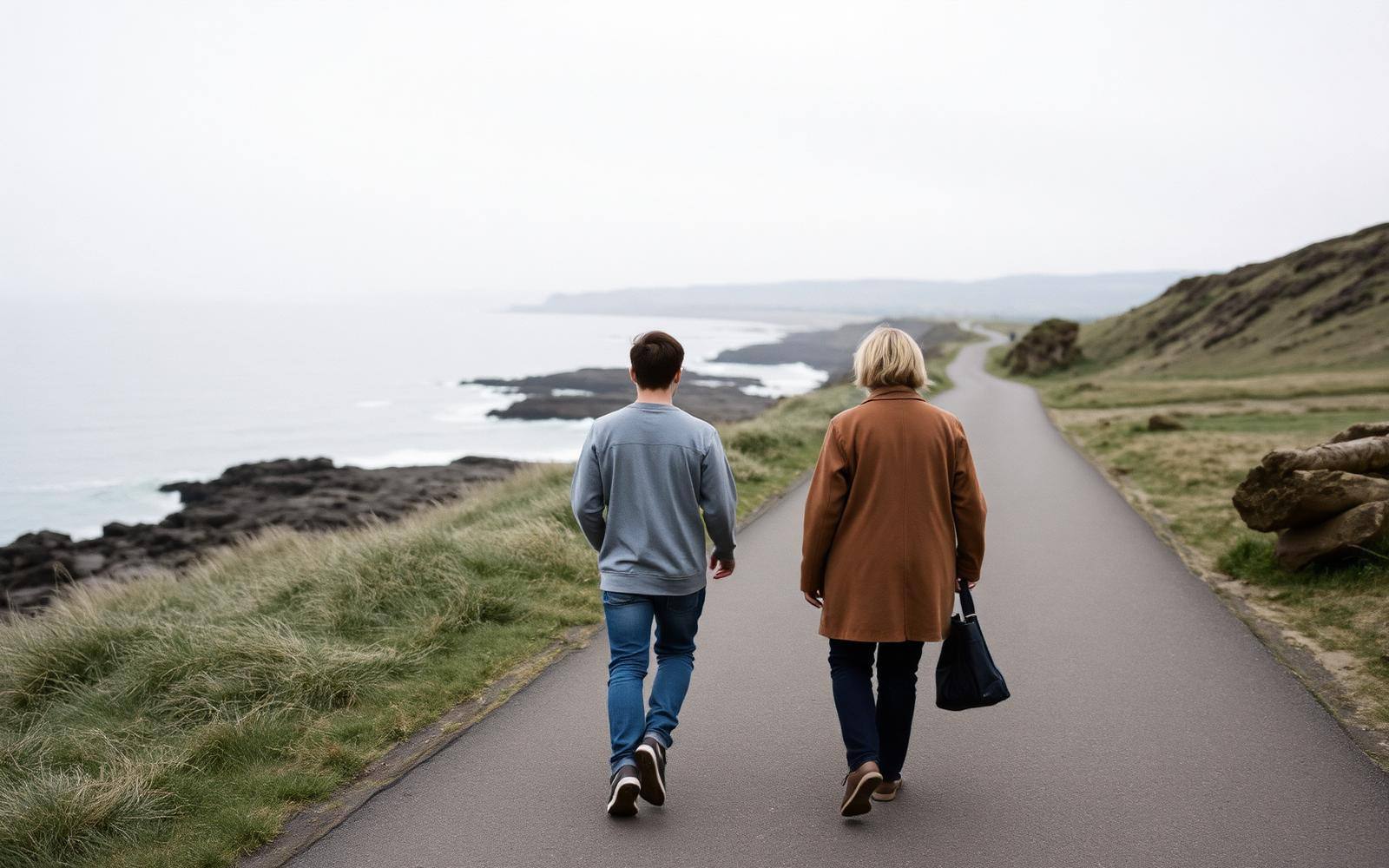 A parent and a teenager walking side by side along a coastal path on an overcast day