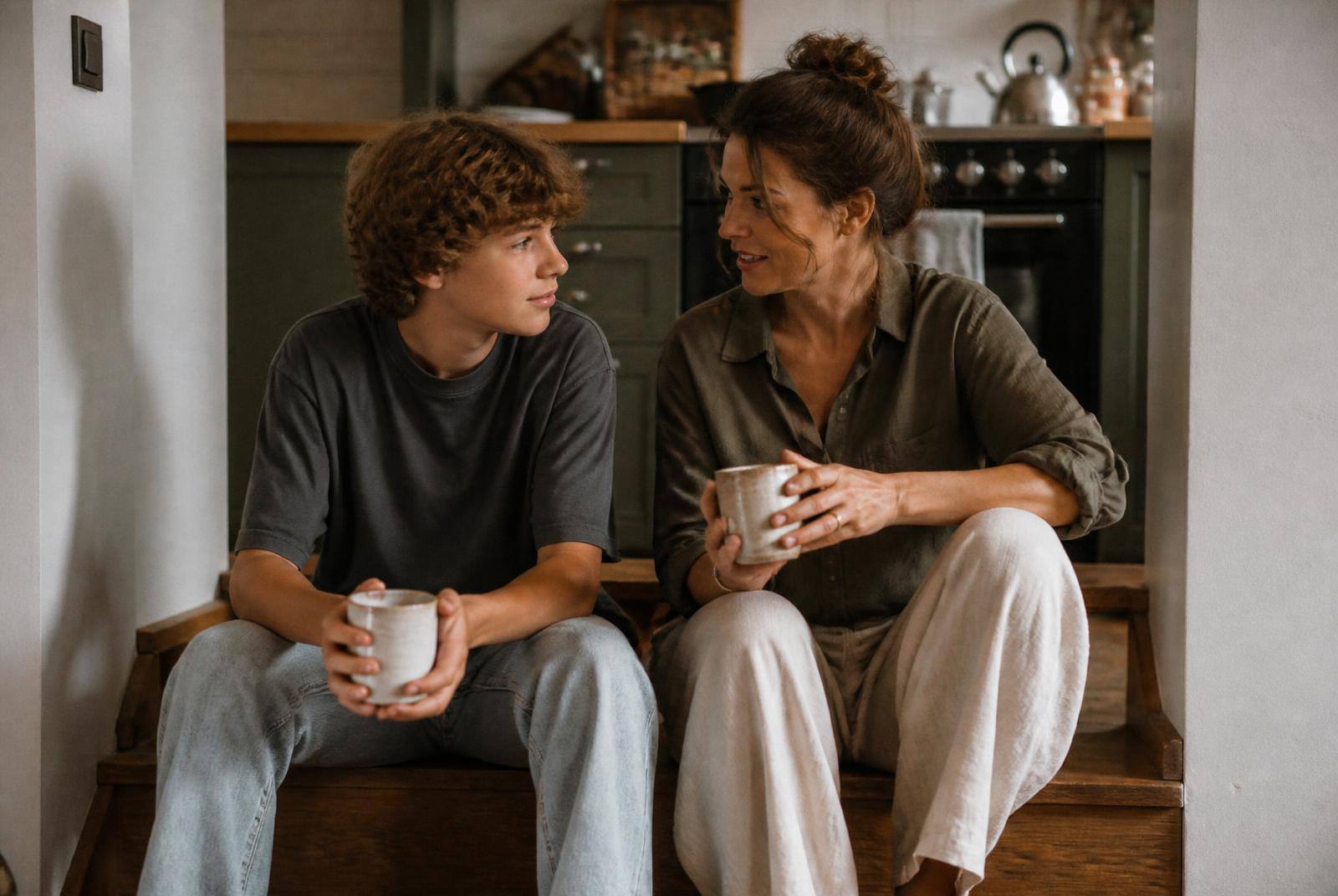 A teenager and a parent sitting side by side on kitchen steps with mugs, mid-conversation