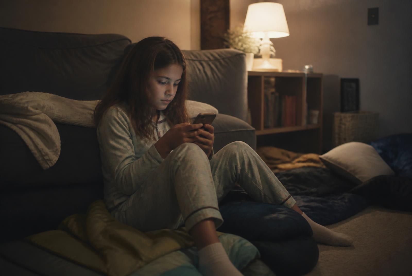A ten-year-old girl in pyjamas sitting on a sleeping bag in a dimly lit living room, holding a phone, sleepover bedding around her