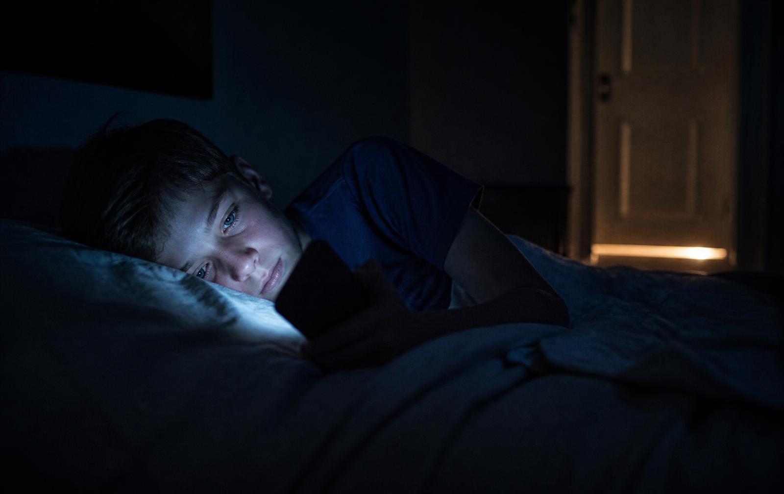 A young teenager lying in a dark bedroom, face lit by the blue glow of a phone screen