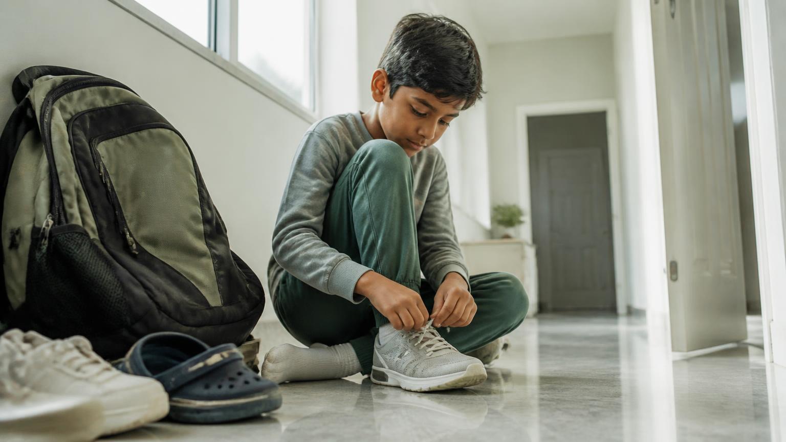 A boy sitting on a hallway floor tying his shoelaces beside a school backpack