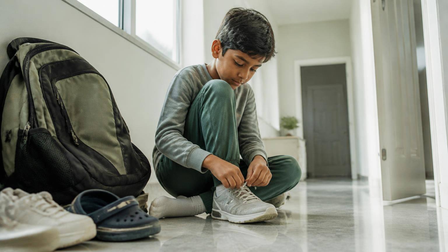 A boy sitting on a hallway floor tying his shoelaces beside a school backpack