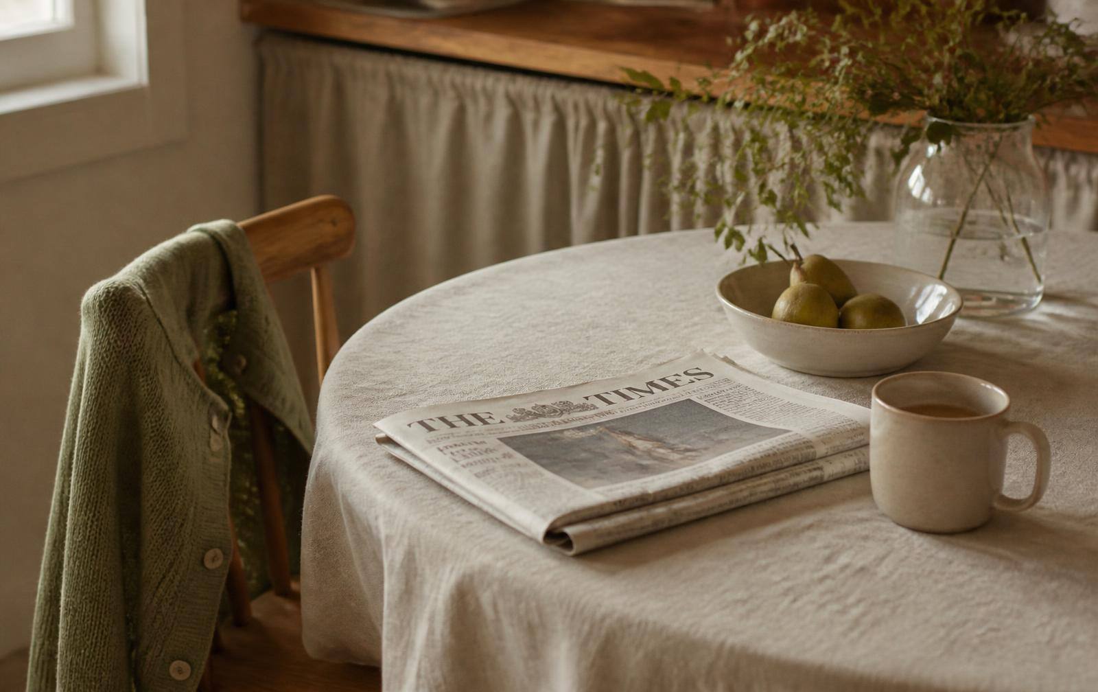 A folded newspaper and a warm mug on a linen-covered kitchen table beside a child's sage cardigan on a wooden chair in morning light