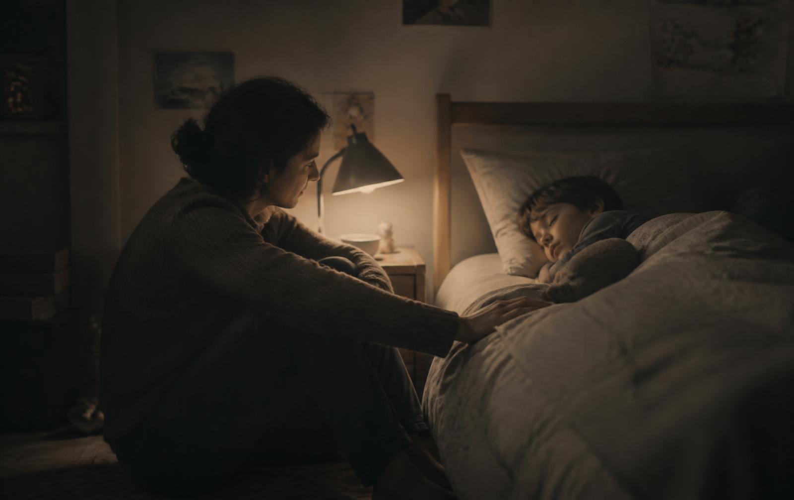 A parent sitting quietly beside a school-aged child's bed in a softly lit bedroom after a frightening night-time awakening
