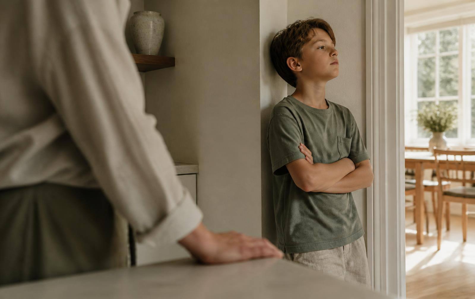 A boy of about ten standing with folded arms in a kitchen doorway, looking away, with a parent's hand resting calmly on the counter nearby
