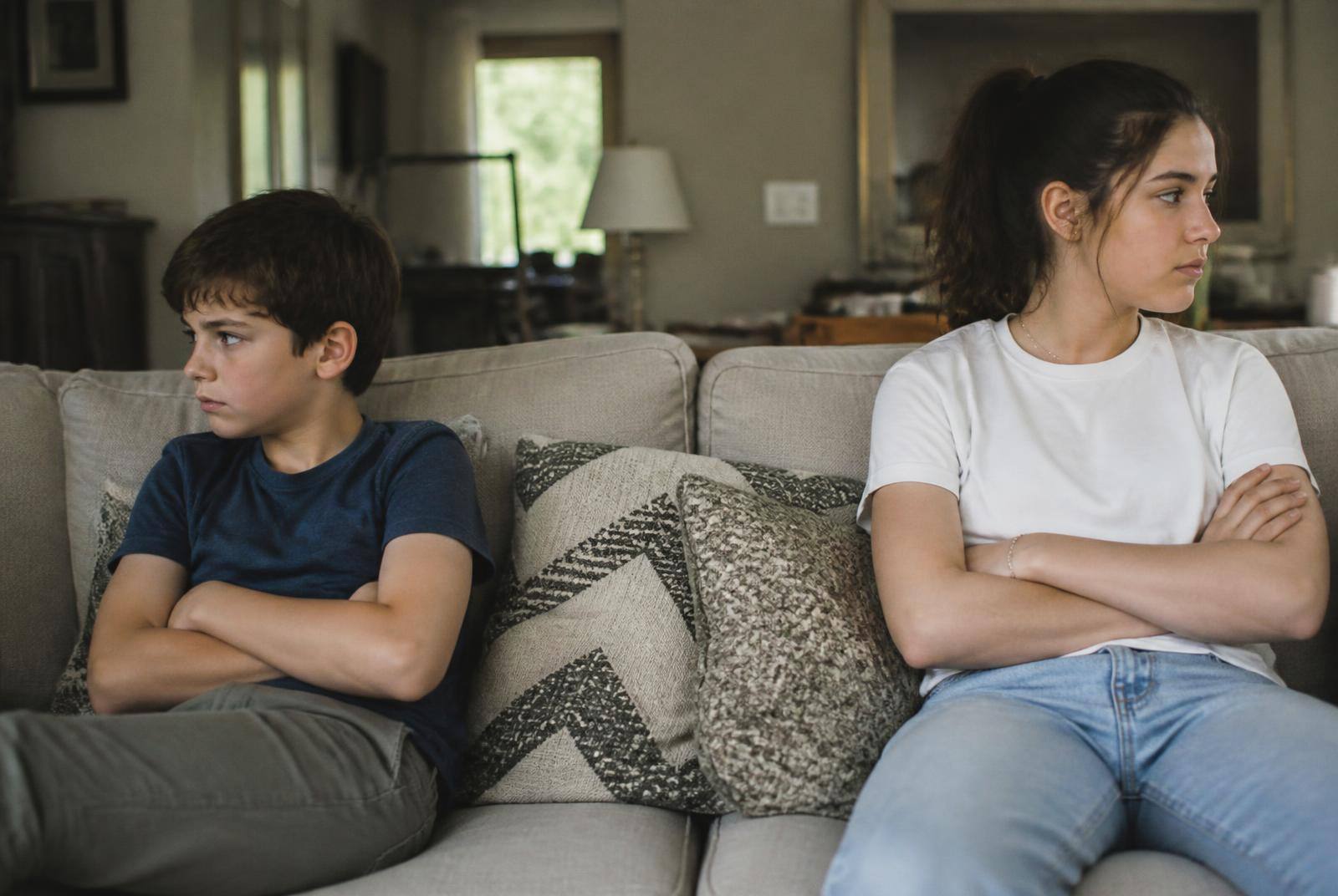 A nine-year-old and a thirteen-year-old sitting apart with folded arms on the sofa after an argument