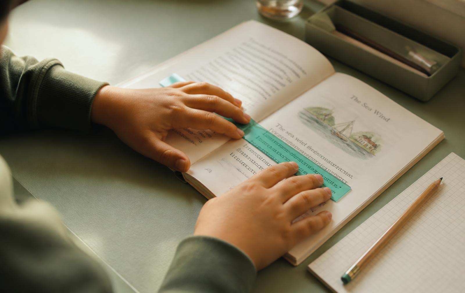 A child's hands tracking a line of text in an open book with a coloured reading ruler