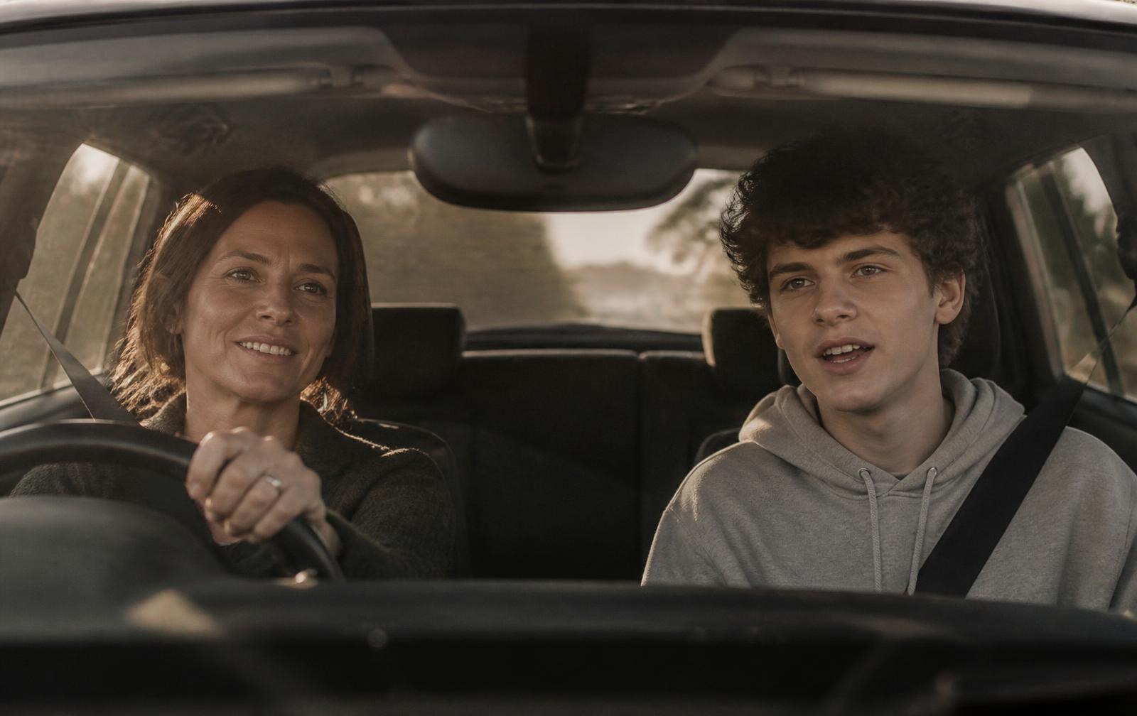 A mother driving with her sixteen-year-old son in the passenger seat, mid-conversation in warm afternoon light