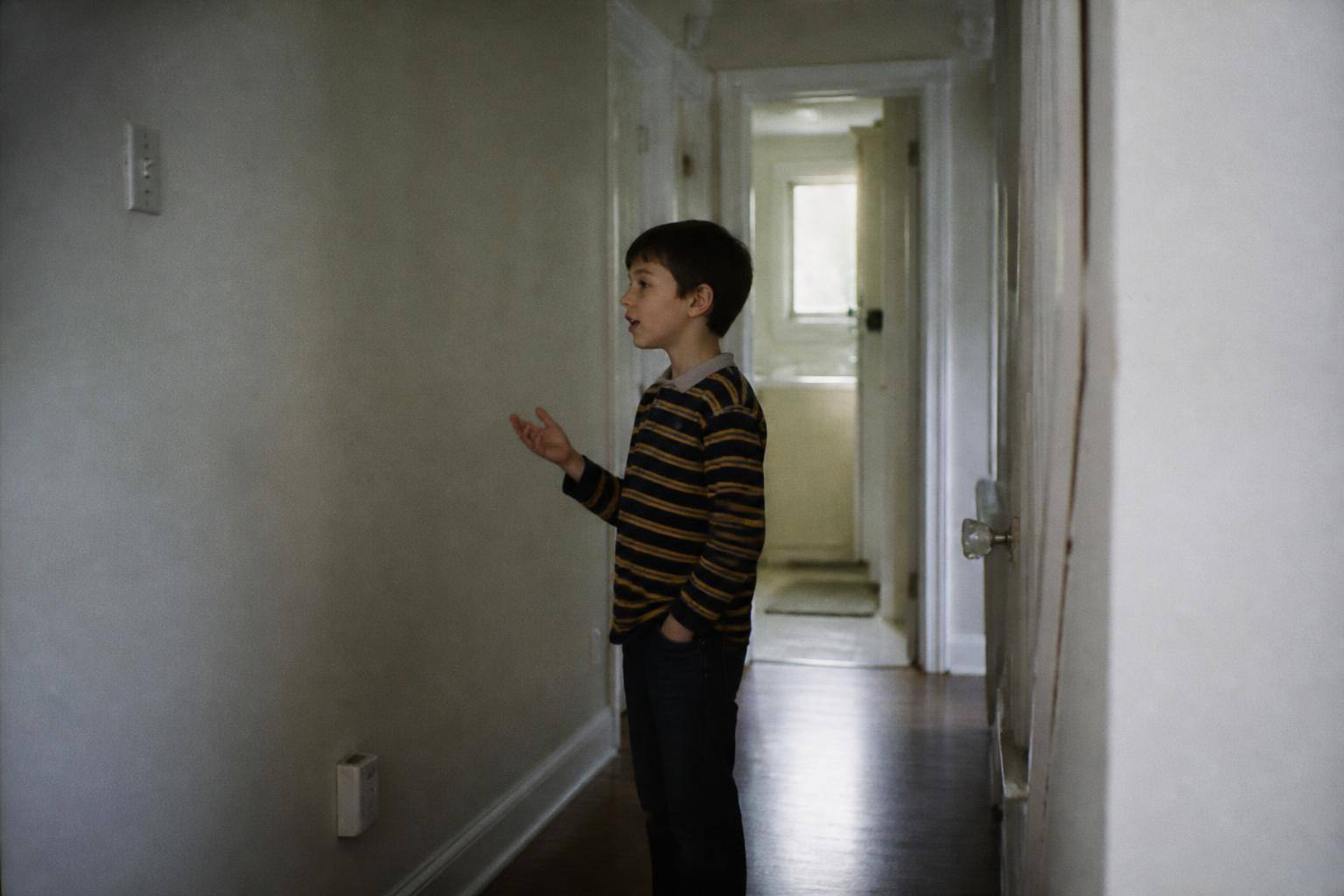 An eight-year-old boy in a bright hallway holding up a calm hand while speaking to someone off camera