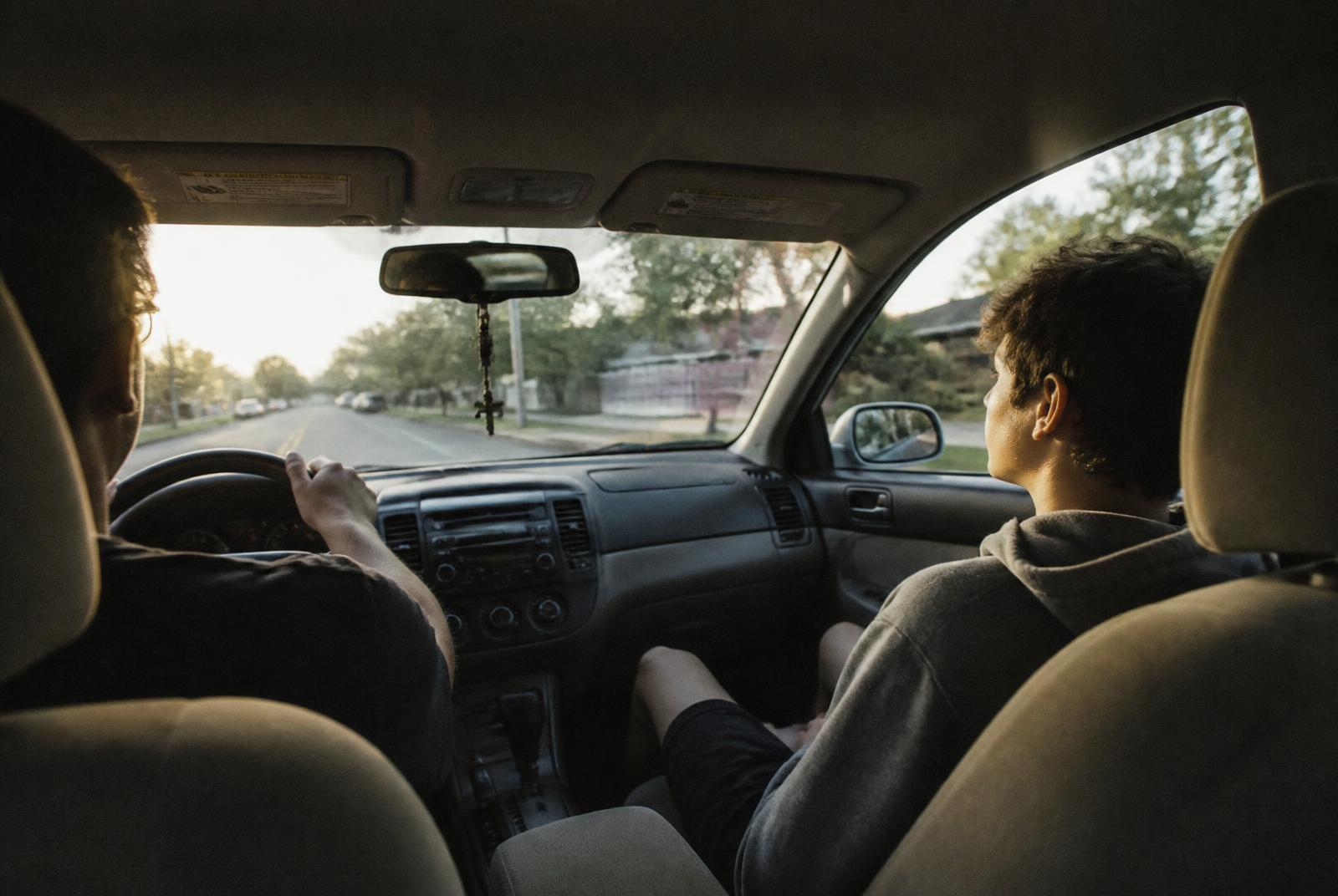 A parent driving with a fourteen-year-old in the passenger seat looking out of the window, late afternoon light on a suburban road