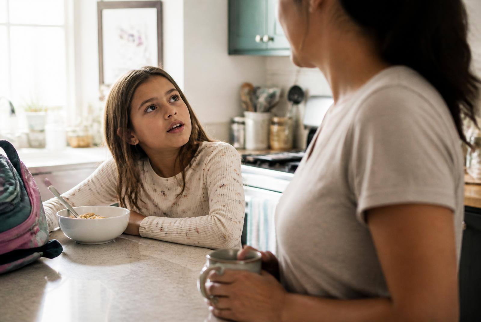 An eight-year-old girl at a kitchen counter looking up at a parent mid-question, morning light, cereal bowl and school bag nearby