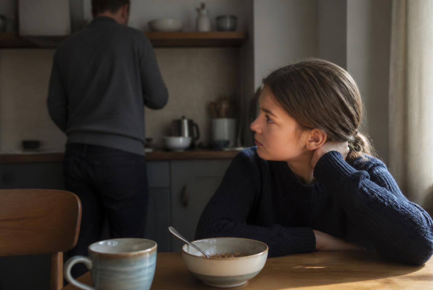 A child at a kitchen table quietly watching a parent's back across the room in morning light