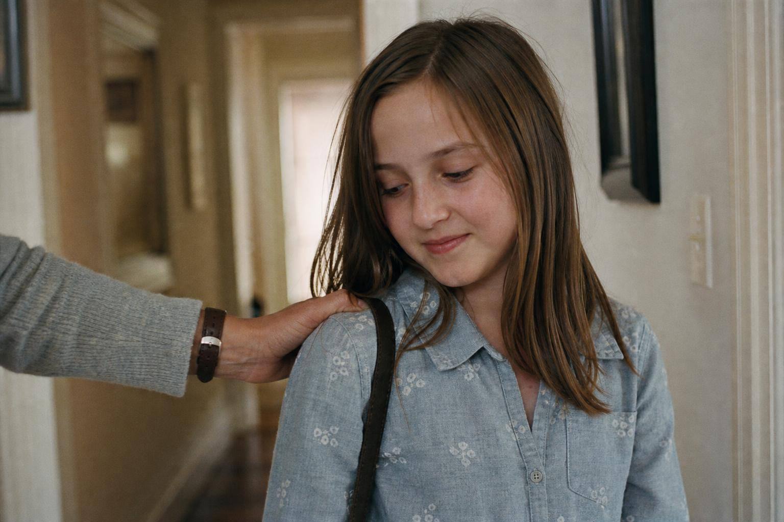 An eleven-year-old girl in a hallway looking down with a small self-conscious smile as a parent's hand rests on her shoulder