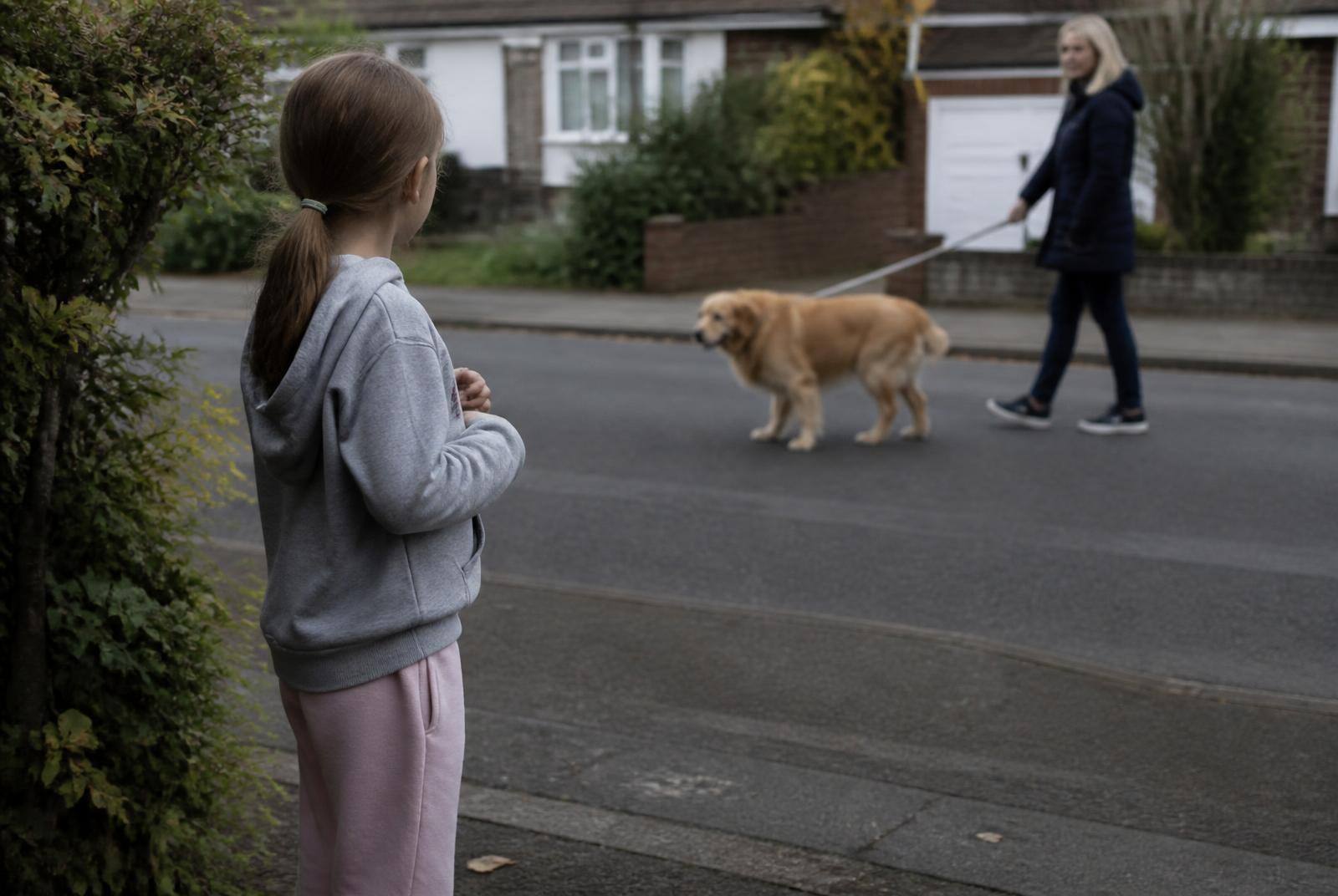 A ten-year-old standing on the pavement watching a dog on a lead across a quiet street