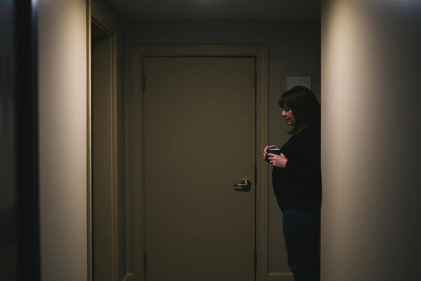 A closed bedroom door in a family hallway with warm light beneath it and a parent standing nearby holding two mugs