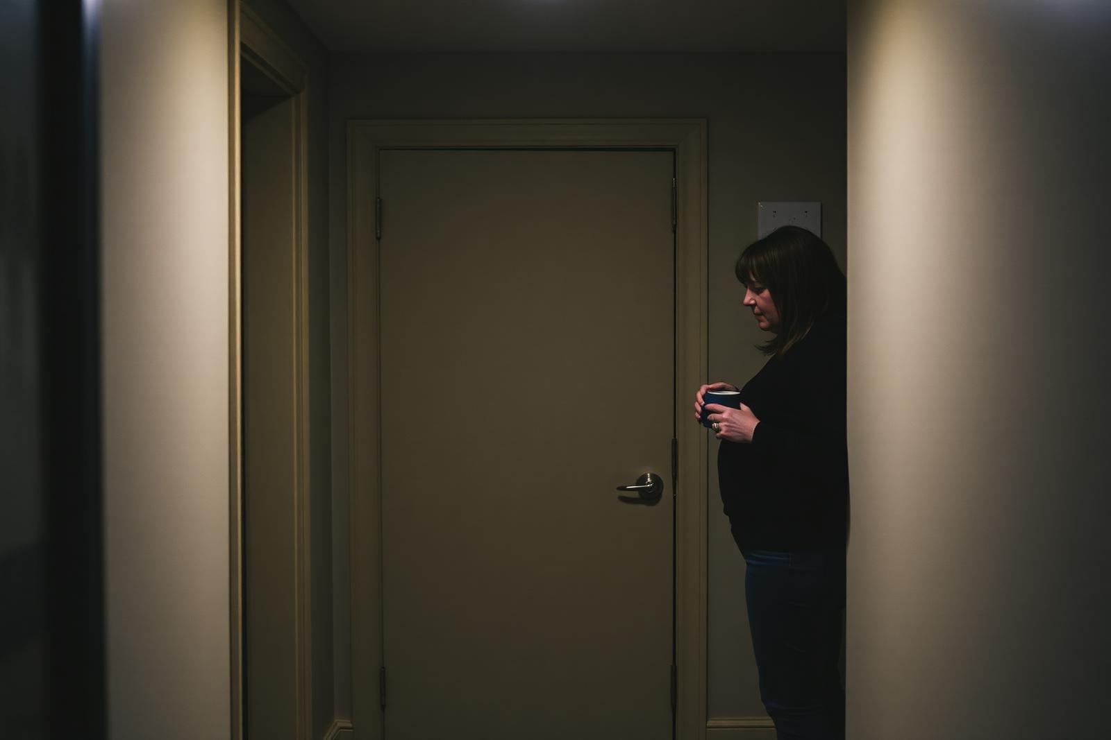 A closed bedroom door in a family hallway with warm light beneath it and a parent standing nearby holding two mugs