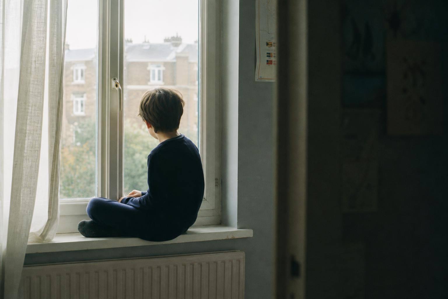 A nine-year-old child sitting alone on a bedroom windowsill looking out at a grey afternoon