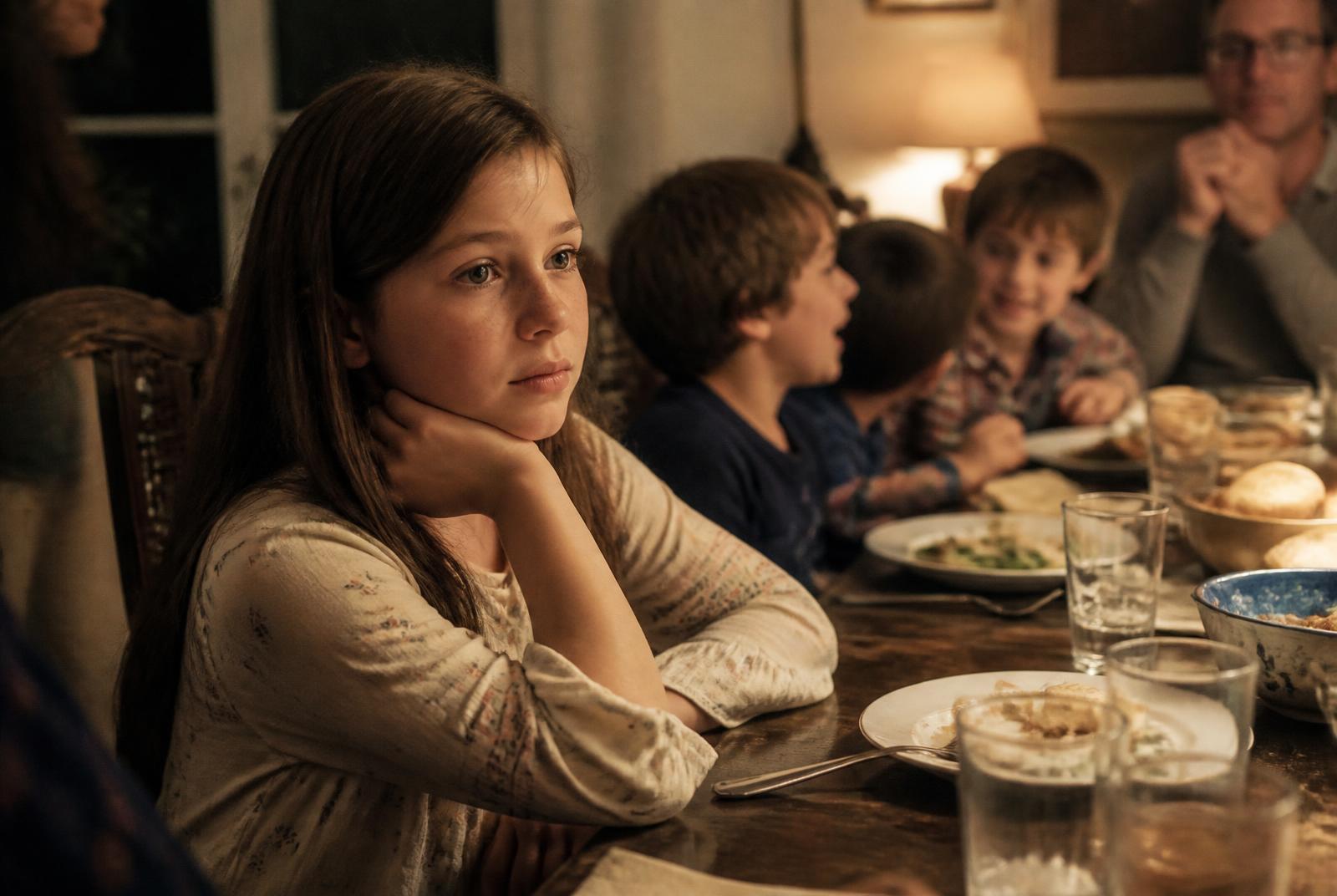 A ten-year-old girl sitting slightly apart at a busy family dinner table, listening, with siblings talking animatedly in soft focus behind her