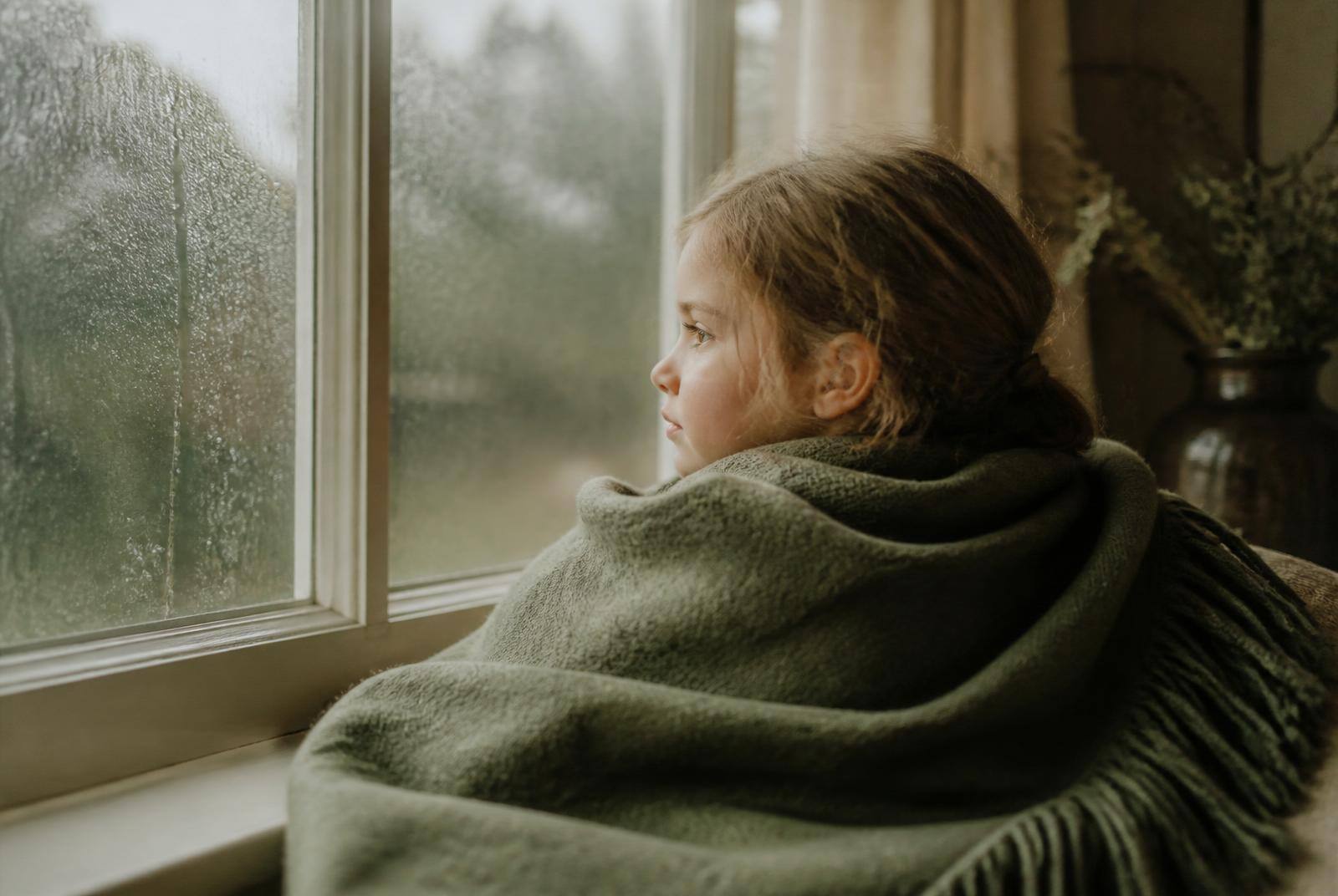 A child wrapped in a green blanket looking out of a rain-streaked window