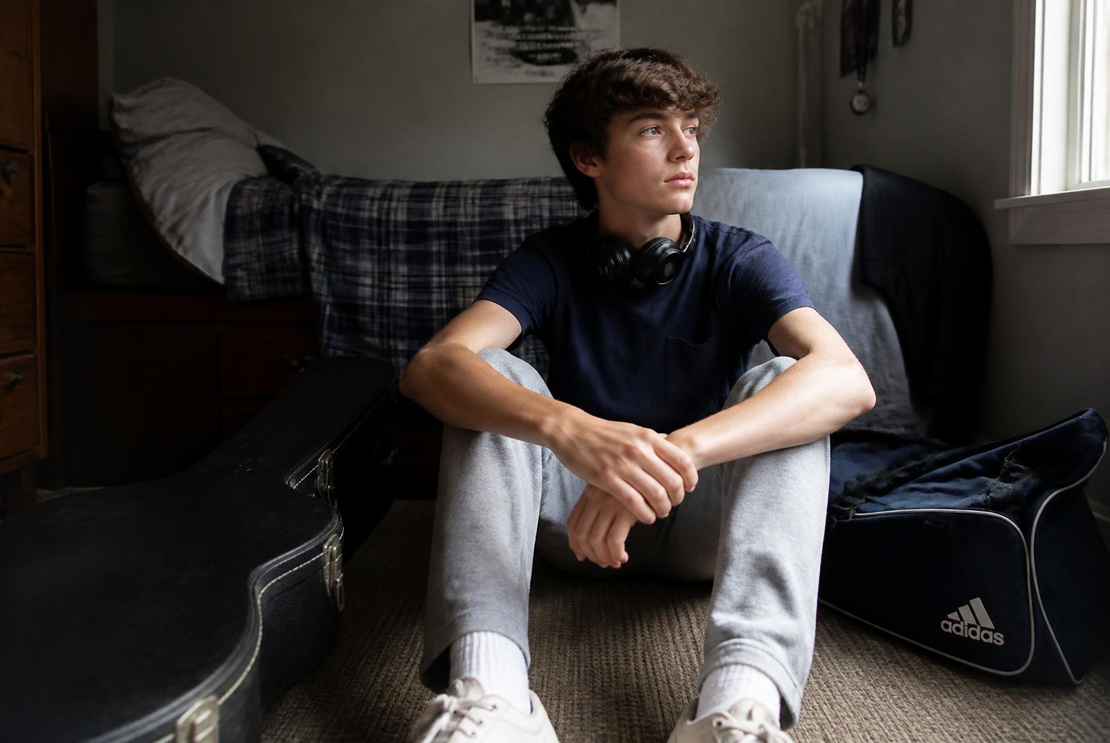 A fifteen-year-old boy sitting on his bedroom floor beside a guitar case and a sports bag, headphones round his neck, late afternoon light
