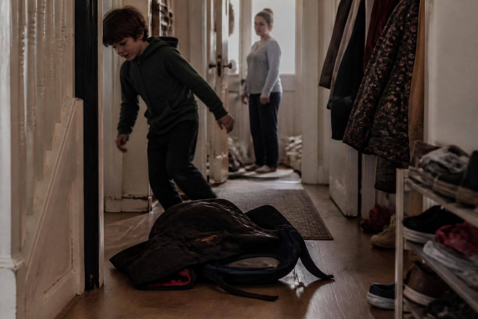A ten-year-old dropping their school bag in the hallway at home while a parent watches from the doorway